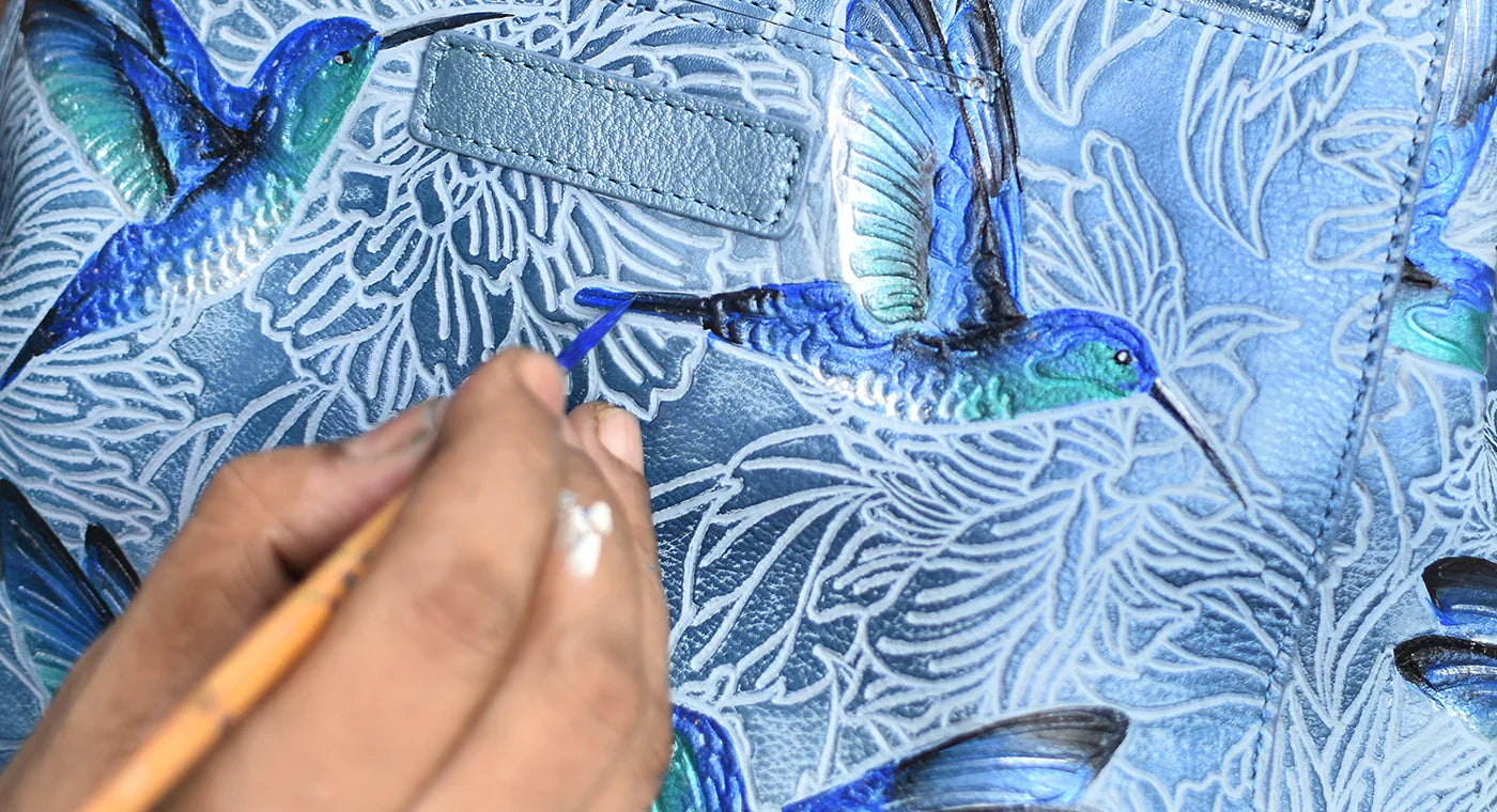 A person paints blue and green hummingbirds on a detailed patterned surface.
