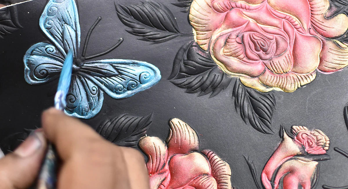 A person's hand using a small brush to paint a detailed embossed design featuring a blue butterfly and colorful roses on a black surface.