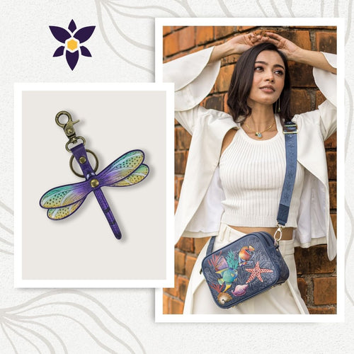 Woman in white outfit carrying a blue Bundle of Twin Top Messenger with Painted Leather Bag Charm - 704-K0034 by Anuschka; inset shows a multicolored dragonfly RFID protected keychain against a neutral background.