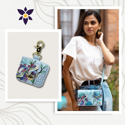 A woman wearing a white blouse is carrying an Anuschka Bundle of 4 in 1 Organizer Crossbody with Airpod Pro Case - 711-1179. An inset shows a close-up of a matching keychain holder with a similar floral pattern and gold clasp, alongside an elegant Airpod Pro case.
