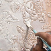 A minimalist artist meticulously paints details on a Two Fold Organizer Wallet - 1178 featuring raised floral and butterfly designs using a paintbrush with white paint by Anuschka.