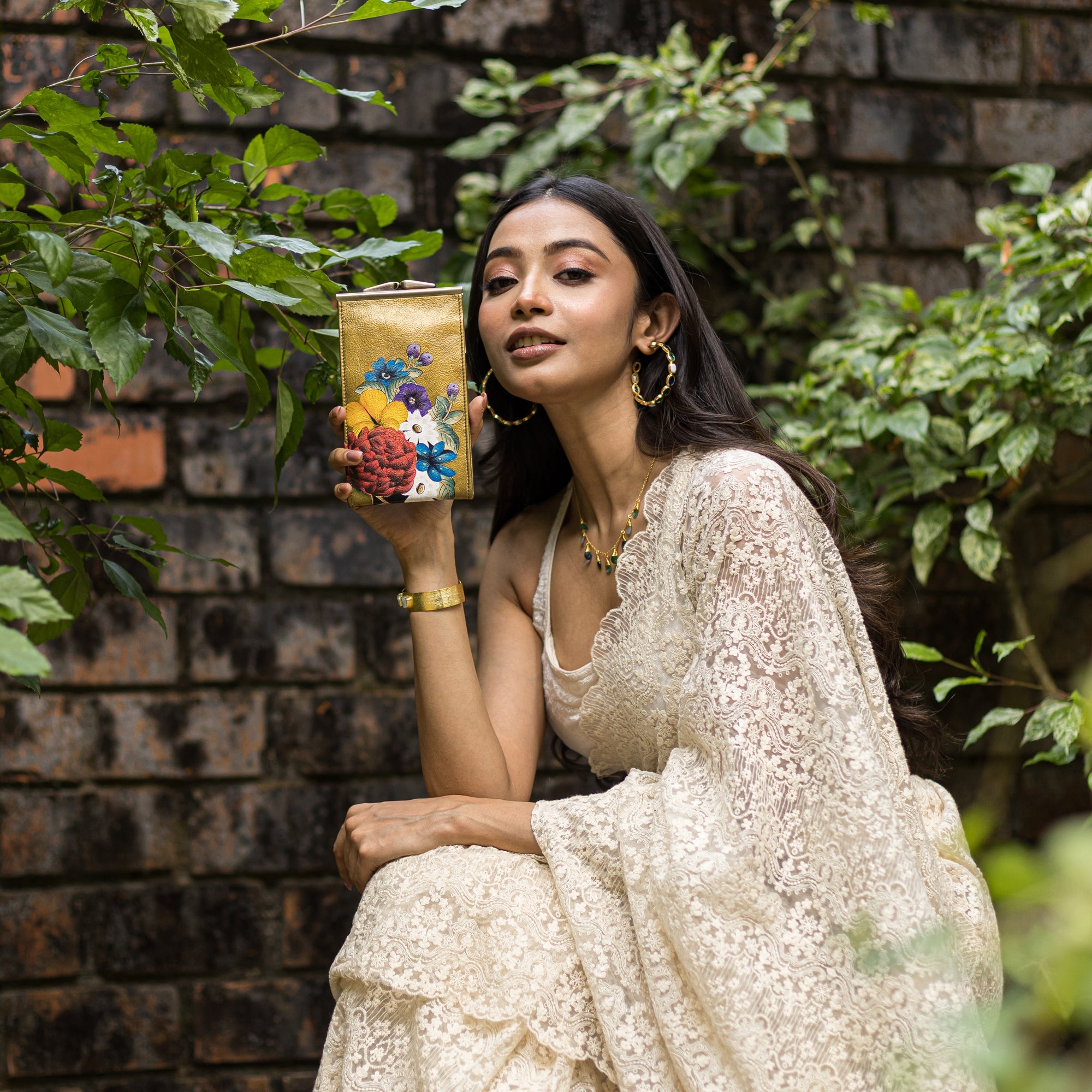 A woman in a white lace saree holds an ornate vase with floral designs against a brick wall backdrop with green foliage, while beside her rests an Anuschka Double Eyeglass Case - 1009.
