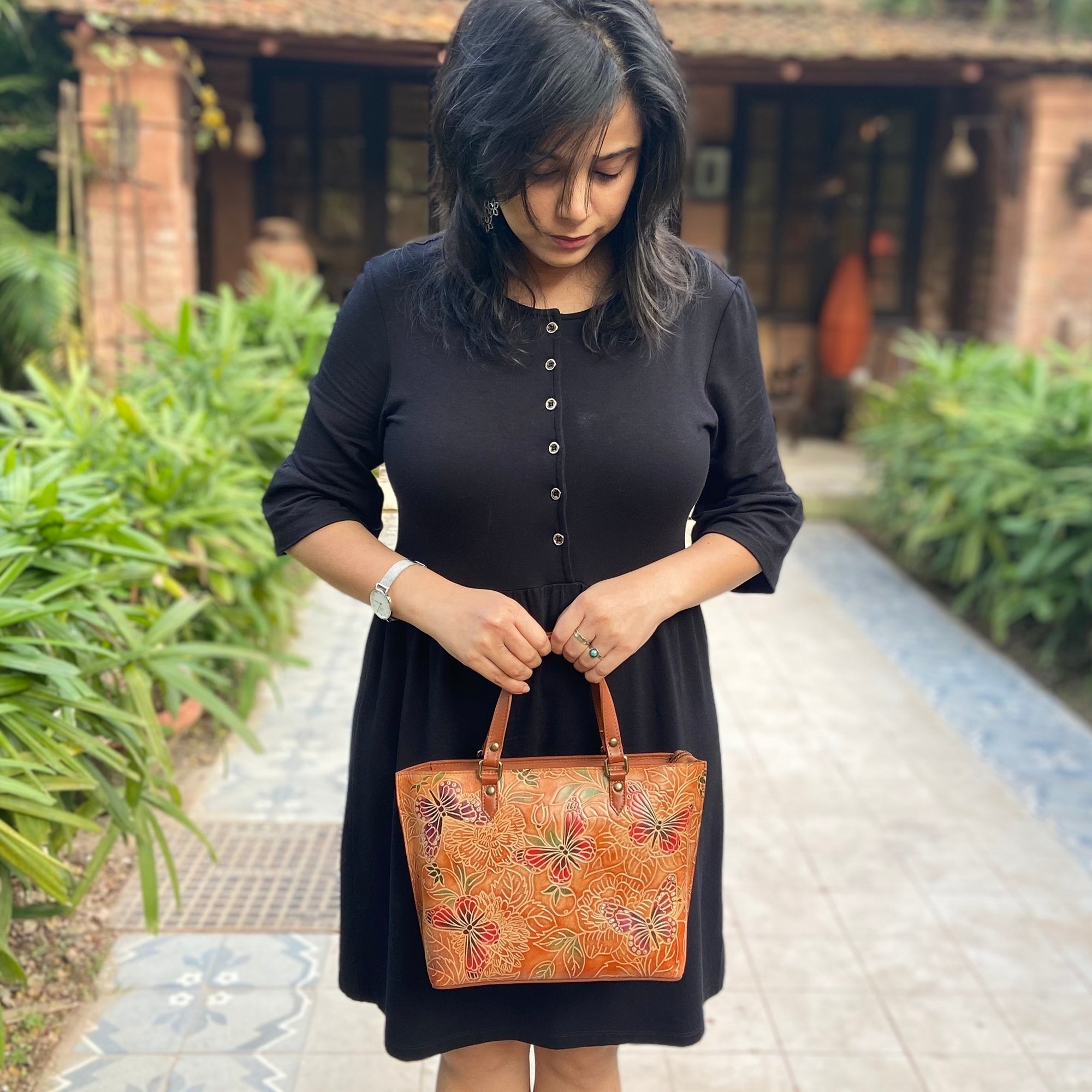 Woman in a black dress holding an ornate, Anuschka Medium Tote - 693, looking down.