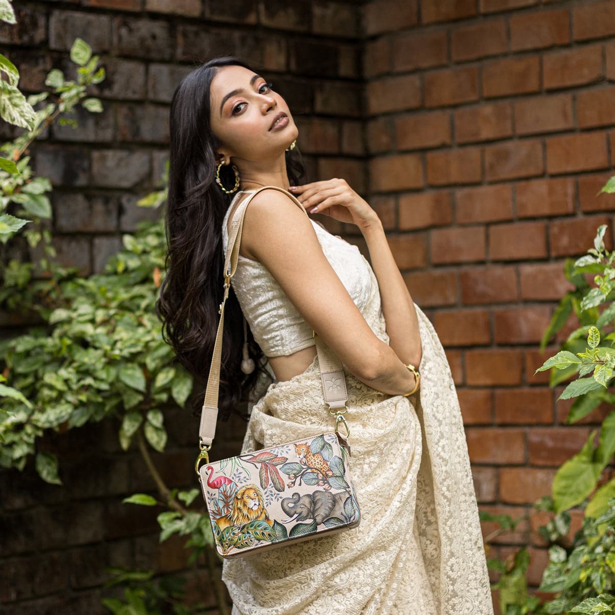Woman traveling light and organized, posing with an Anuschka Triple Compartment Crossbody-696 against a brick wall with green foliage.