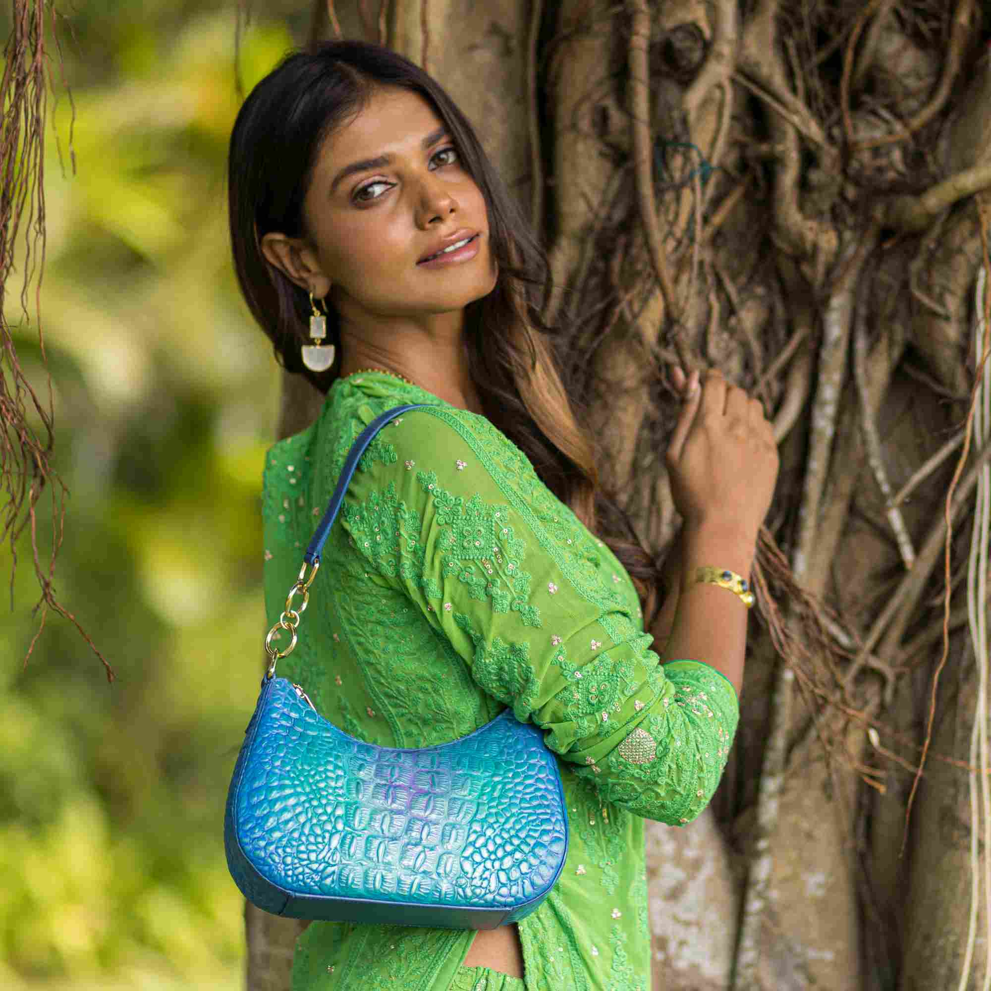 Woman in a green outfit holding a blue Anuschka Small Convertible Hobo - 701 with a crossbody strap, standing next to a tree.