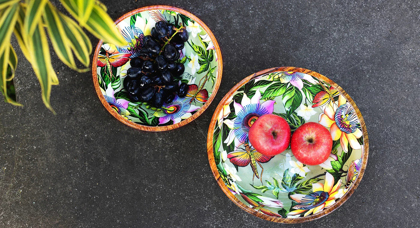 Two colorful bowls on a dark surface; the left bowl contains dark grapes, and the right bowl has two red apples. A plant with green and yellow leaves is partially visible on the top left corner.