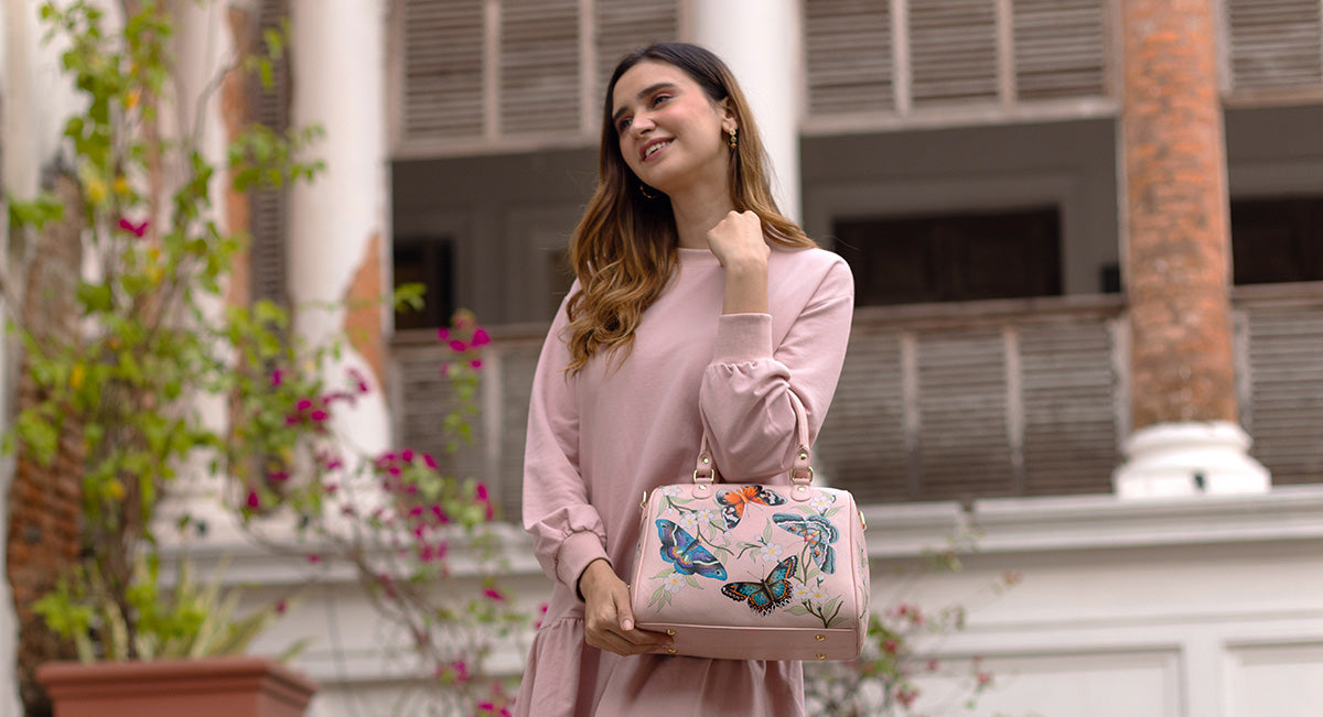 A woman in a pink dress and holding a handbag with butterfly designs stands smiling in front of a building with columns and balconies.
