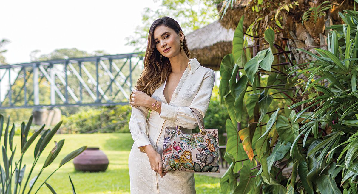 A woman in a white dress holds a colorful tote bag, standing in a lush garden with trees, plants, and a bridge in the background.