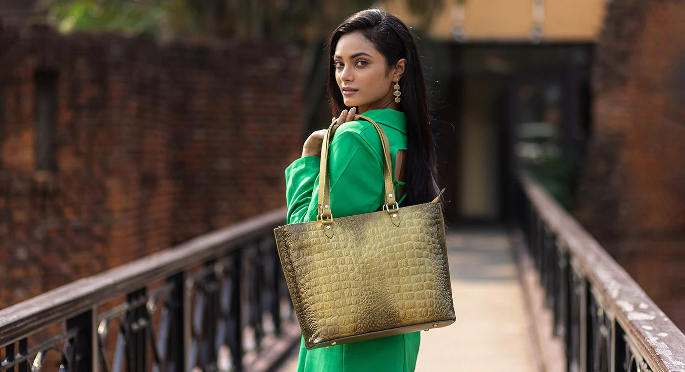 A woman with long dark hair stands on a bridge wearing a green outfit and holding a gold handbag over her shoulder, looking back at the camera.