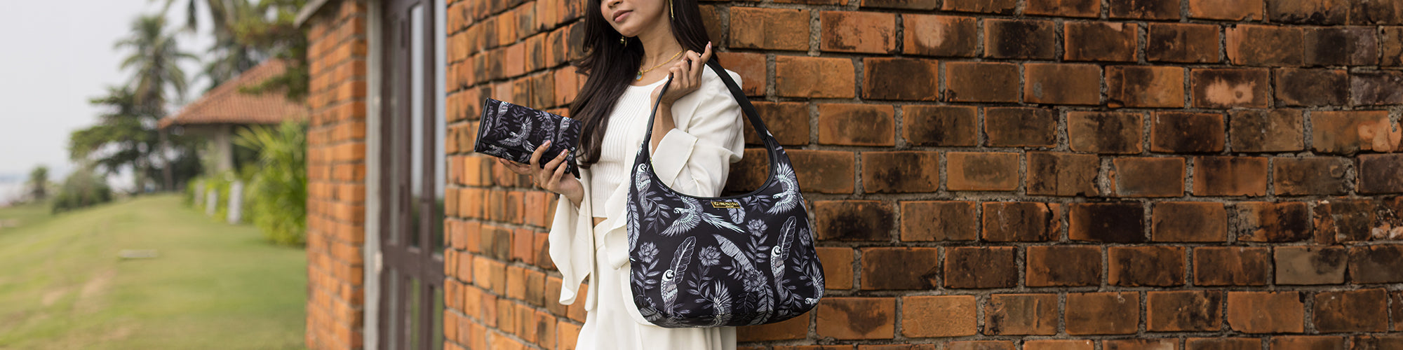 A woman in a white outfit poses next to a brick wall, holding a black and white floral-patterned bag and clutch.