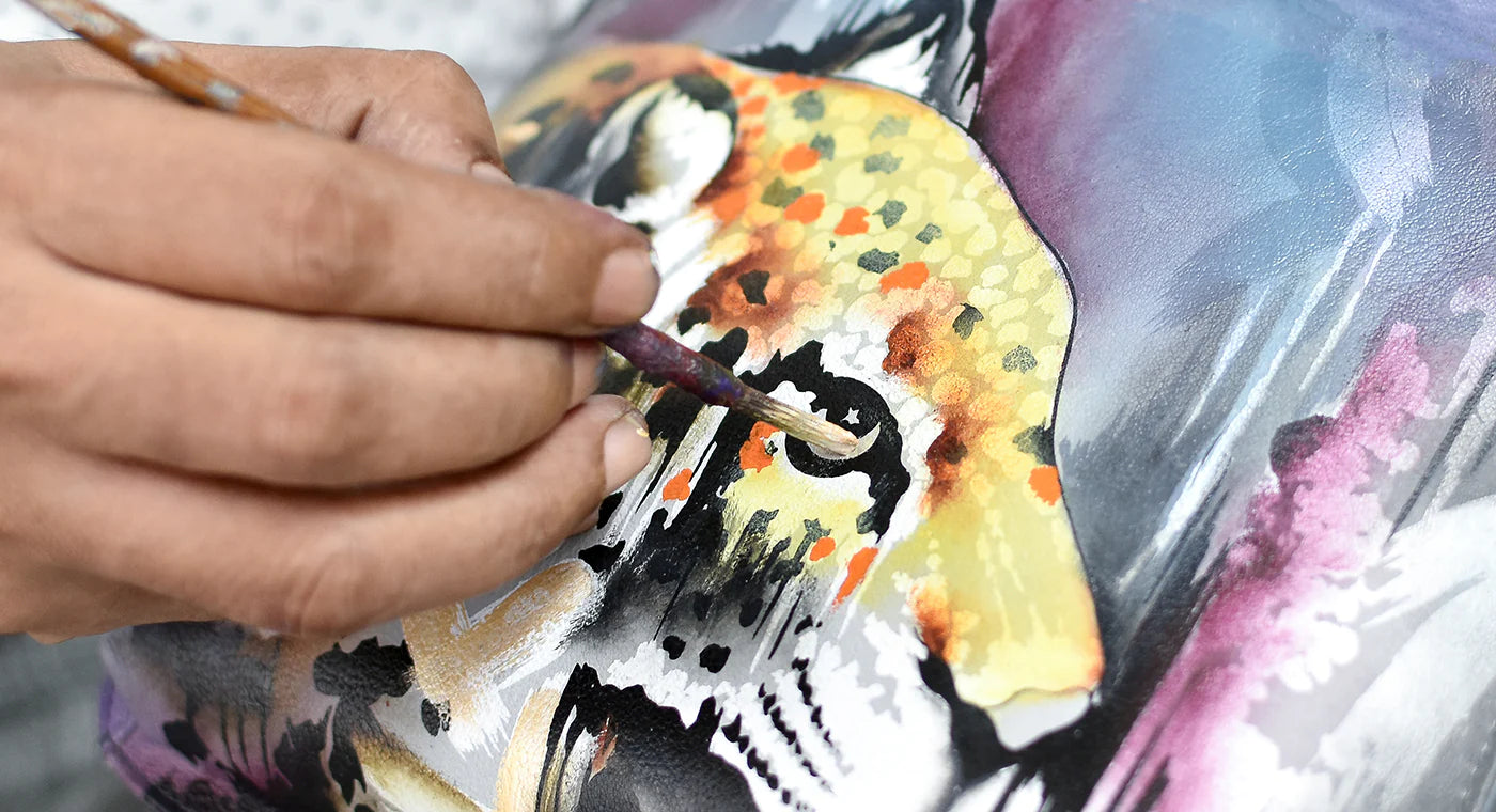 A close-up of a hand painting a colorful, abstract fish design on canvas with a brush.