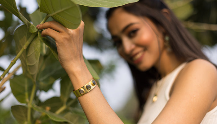 A woman with long hair holds a leaf while smiling. She is wearing a white sleeveless top and a gold bracelet.