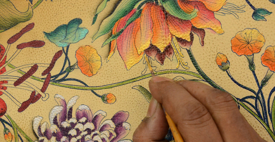 A person's hand holds a brush and paints intricate floral designs in bright colors on a yellow surface.