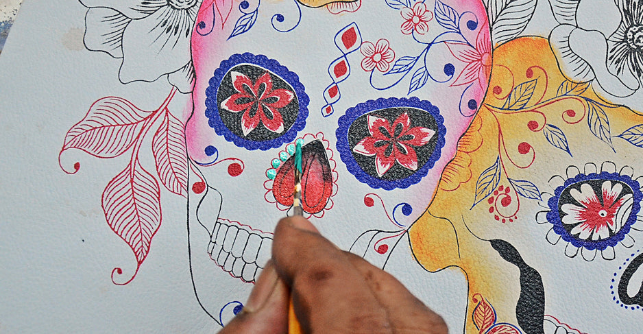 A person is painting a colorful, floral-decorated skull design on paper using a small brush.