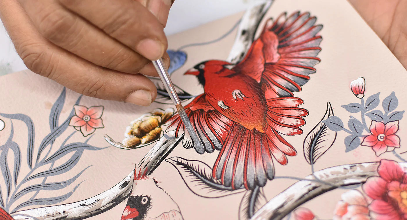 A hand is painting a detailed image of red and white birds perched on branches with flowers using a small brush.