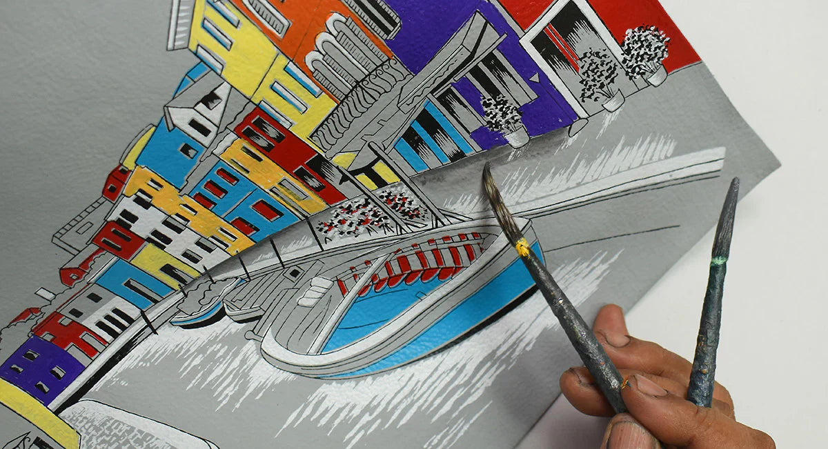 Close-up of hands holding two paintbrushes painting a colorful, abstract cityscape on paper. Brightly colored buildings and a bridge are visible on the artwork.