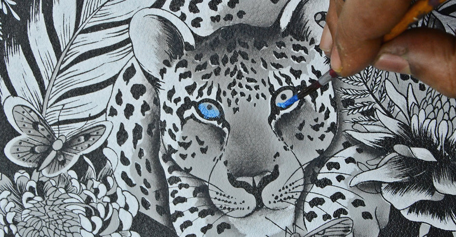 A person is painting the blue eyes of a leopard in a detailed black and white illustration featuring flowers, leaves, and a butterfly.