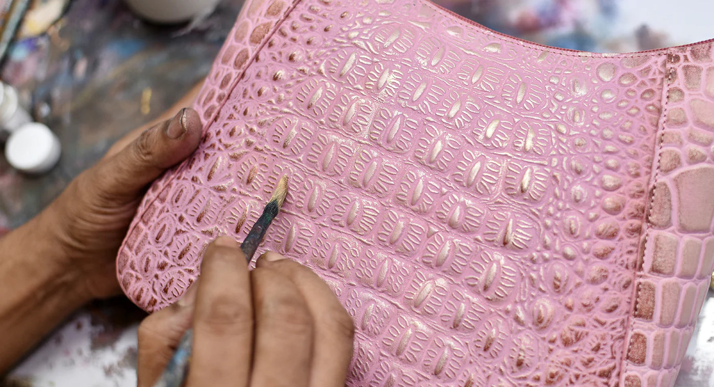 Hands of a craftsperson applying detailed carvings on a pink leather surface with tools and paint around.