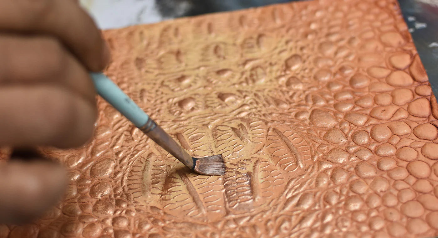 A hand applies paint with a brush to a textured leather surface with a reptile-like pattern.