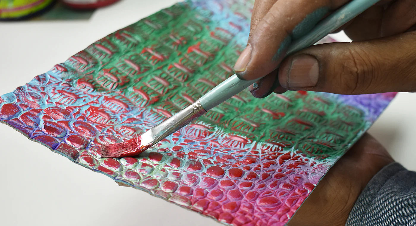 A person using a brush to paint on a textured canvas with iridescent colors and raised stenciled letters.