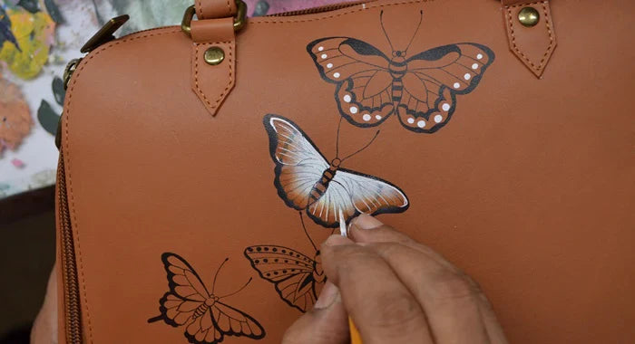 A person paints intricate butterfly designs on a brown leather bag using a fine brush.