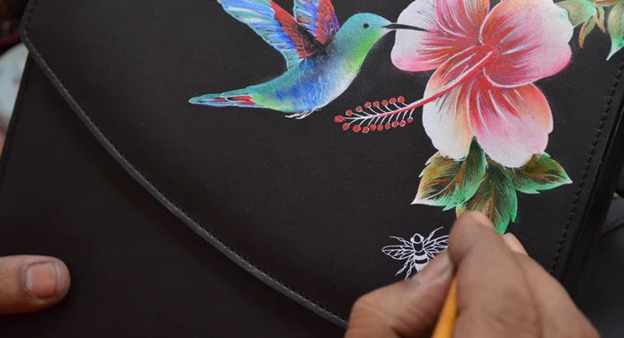 Close-up of a hand painting a hummingbird and a flower on a black leather surface.