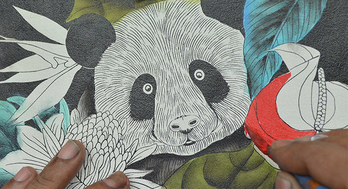 A close-up of hands coloring an illustration of a panda surrounded by flowers and foliage, with some areas already filled in with bright colors.