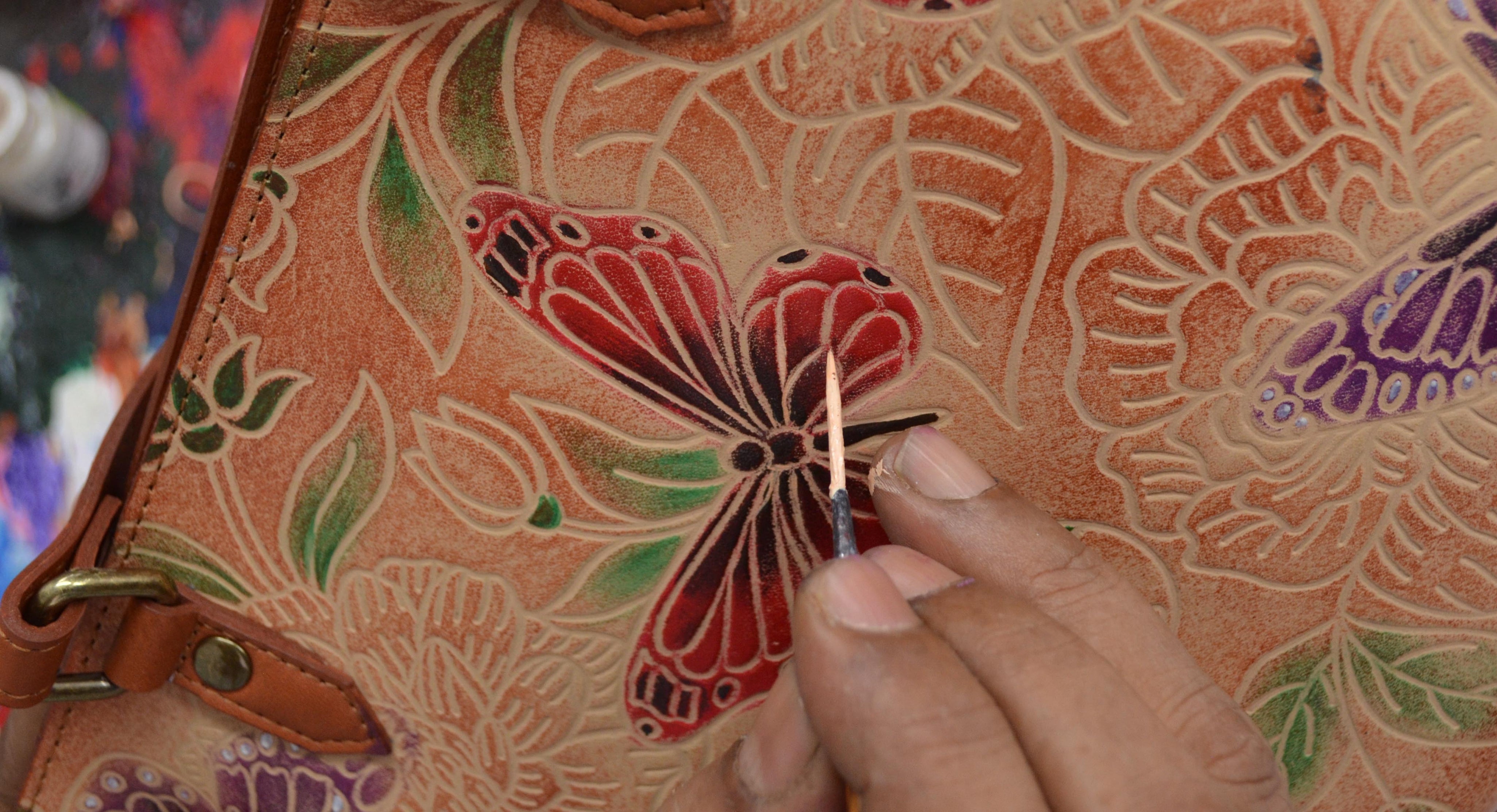 A person uses a small tool to add detail to a colorful butterfly design on a leather surface with floral patterns.