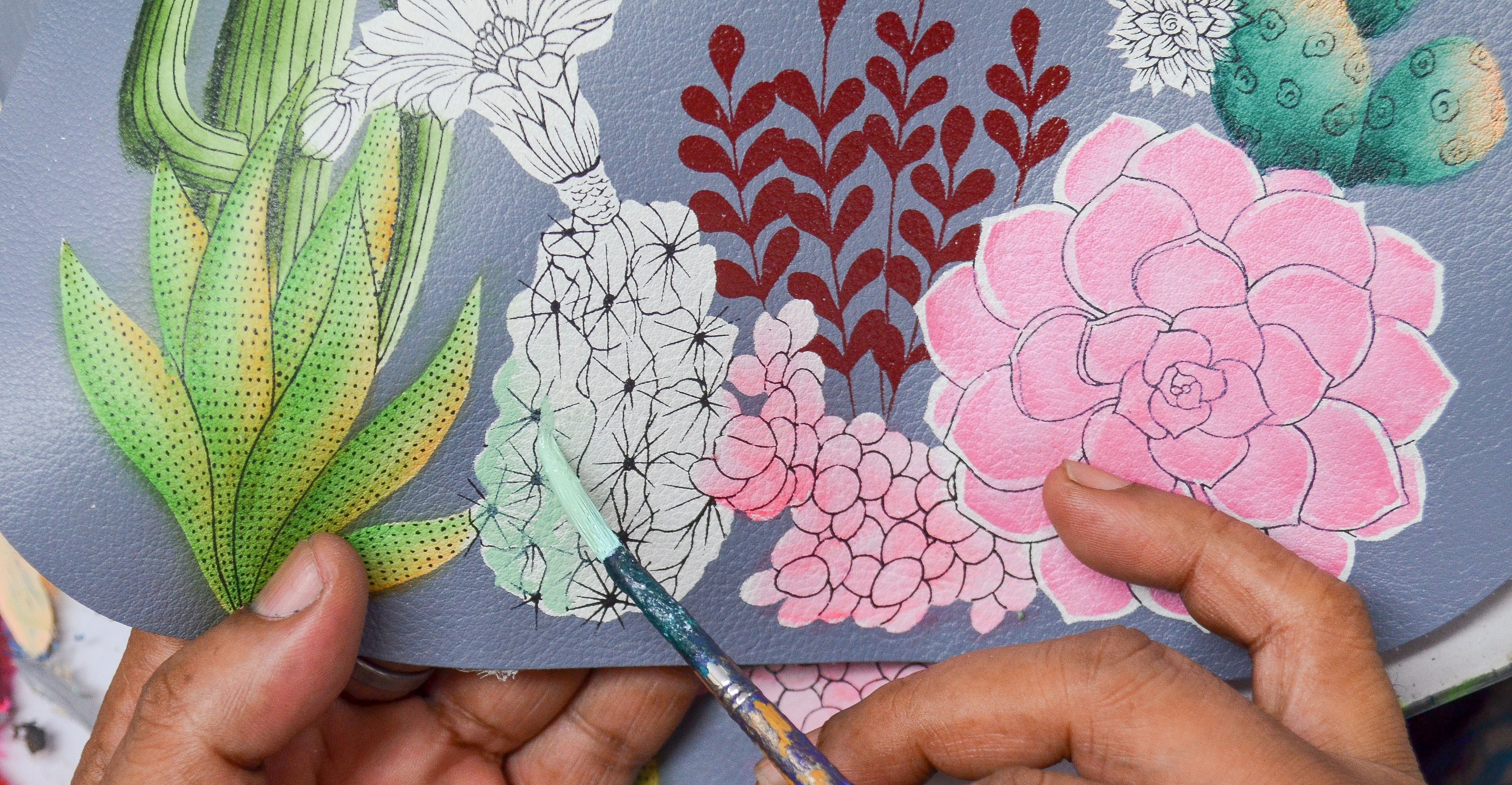 A person painting a detailed floral design with a paintbrush on paper, highlighting pink and green colors.
