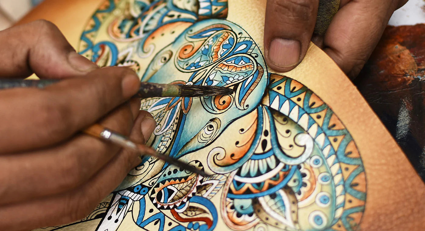 Close-up of hands painting intricate, colorful patterns with fine brushes on a canvas. The design includes various swirling shapes and detailed motifs in blue, orange, and white.