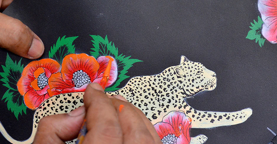 Close-up of a person painting a leopard among vibrant red poppies on a dark background. Hands are visible holding a brush, detailing the artwork.