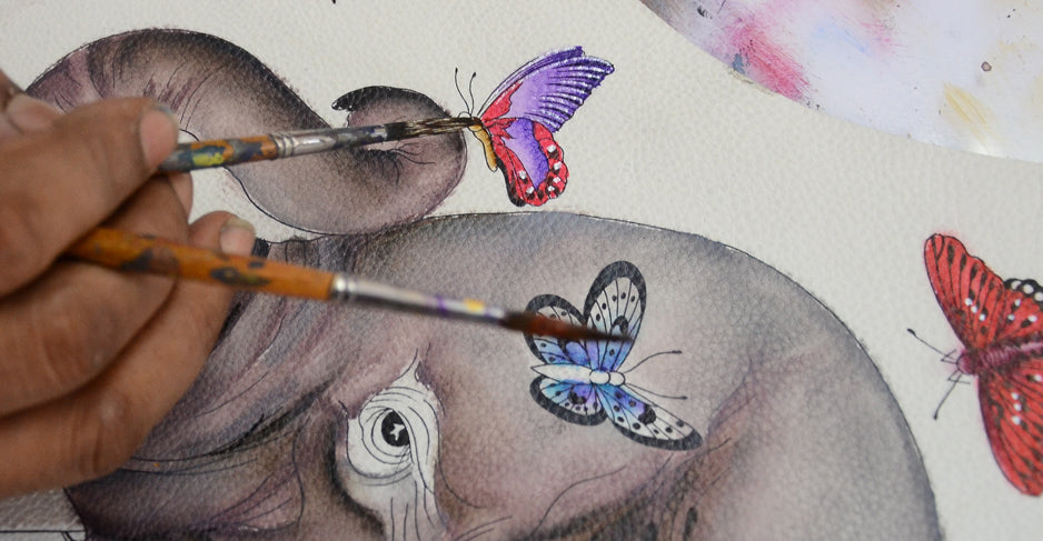 Close-up of an artist's hand painting colorful butterflies on an elephant's face. The brushes apply fine details and bright hues to the artwork.