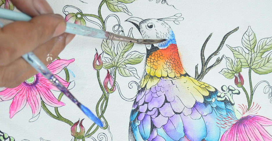 A hand painting a detailed, colorful bird with watercolors. The bird is surrounded by various plants and flowers in the artwork.