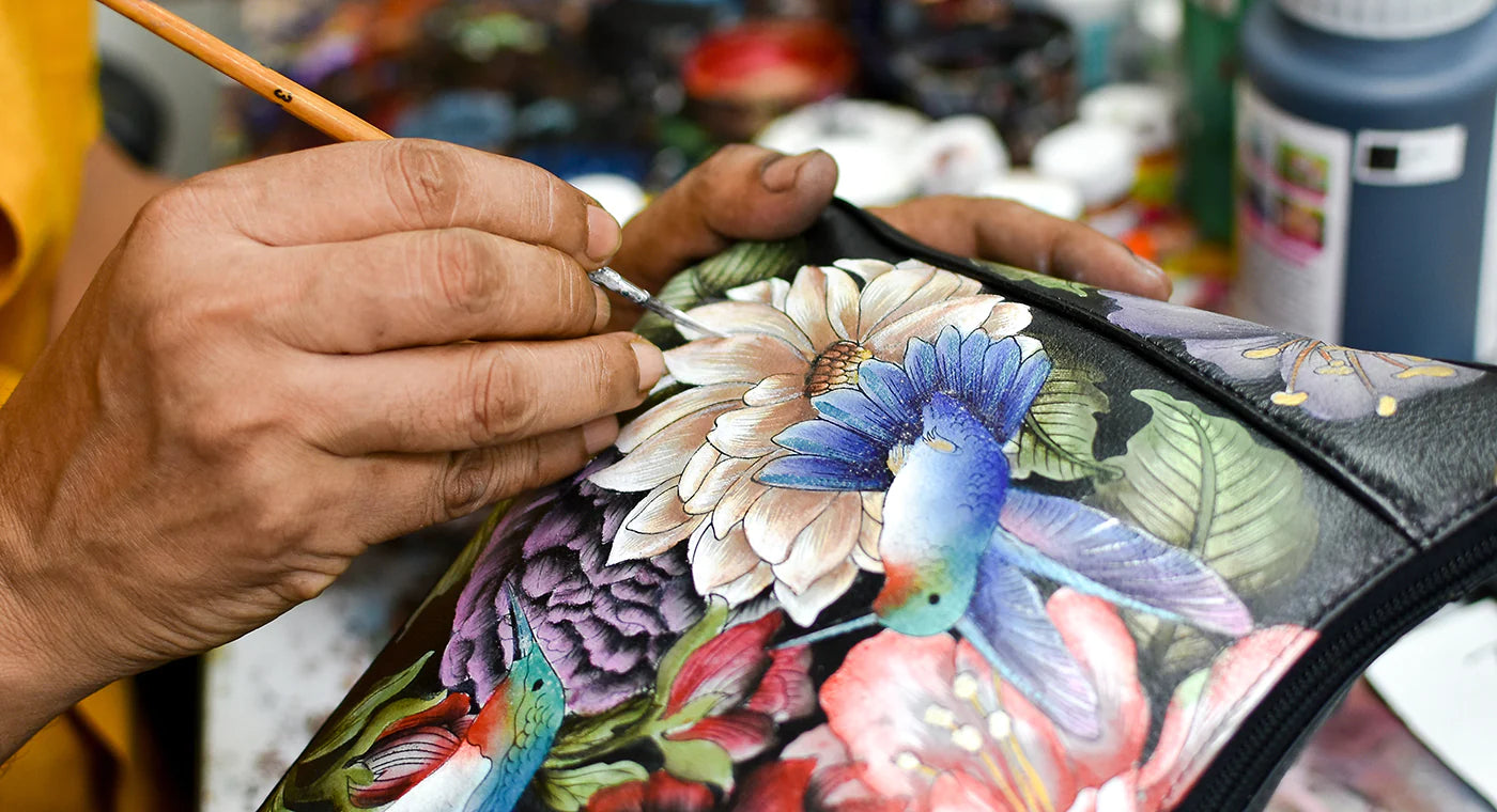 Close-up of a person's hands painting a colorful floral and bird design on a black fabric surface, surrounded by paint bottles and brushes.