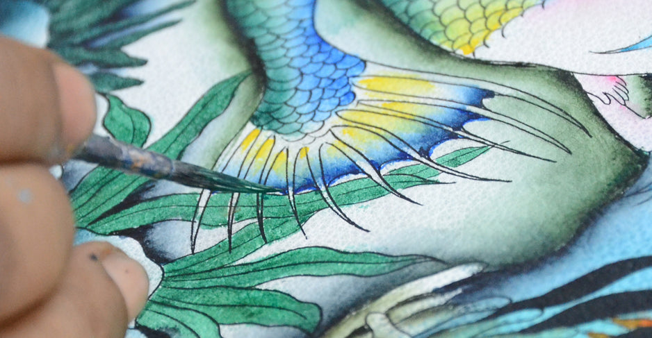 Close-up of a person's hand holding a brush while painting an intricate and colorful fish and plant illustration.