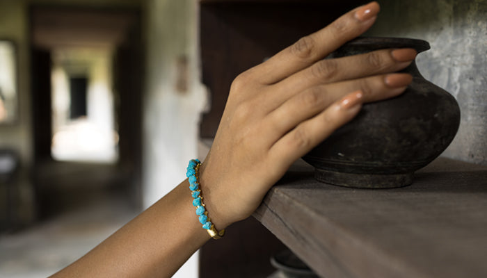 A woman's hand with a turquoise and gold bracelet resting on a wooden ledge next to a black pottery bowl, in a softly lit hallway.