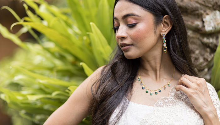 A woman with long dark hair wears a white outfit and poses outdoors with green foliage in the background. She displays matching jewelry, including earrings and a necklace with green gemstones.