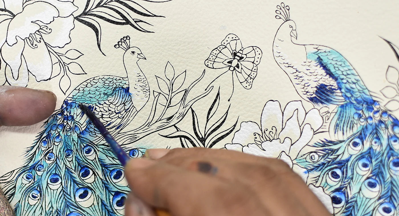 An artist's hand painting detailed blue peacock feathers on a sketch with floral motifs.