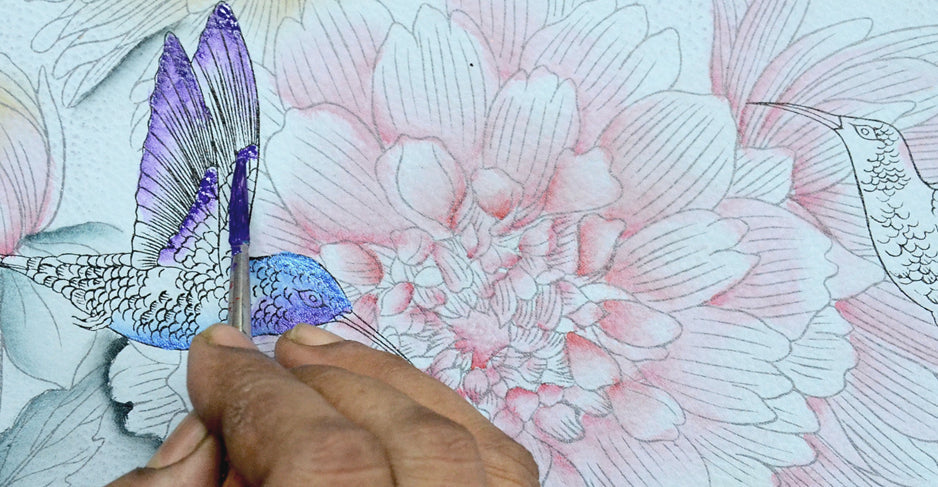 A hand uses a paintbrush to color a detailed flower and hummingbird illustration, adding shades of blue and purple to the bird's feathers.