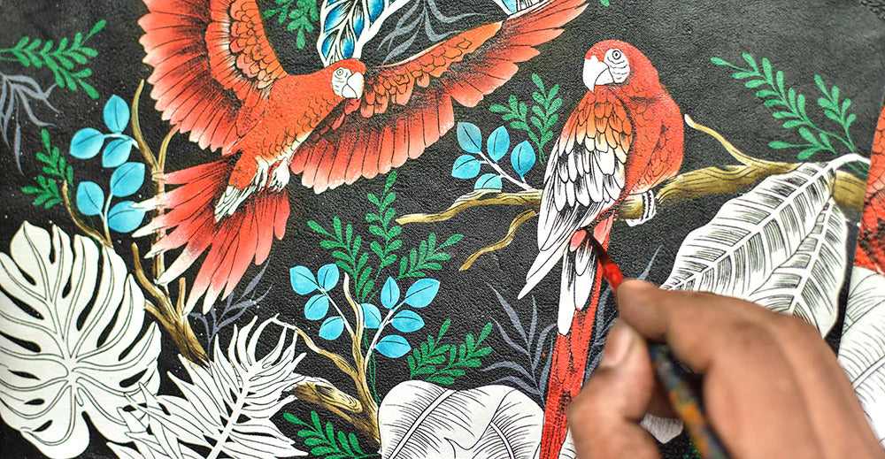 A person is painting a detailed mural of two red parrots surrounded by green and white foliage on a black background.