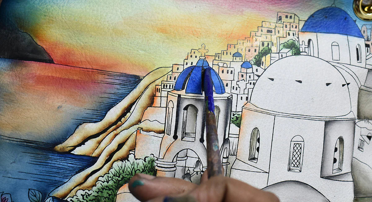 A hand paints blue roofs on a detailed watercolor illustration of a coastal village at sunset, featuring white buildings on a hillside and a colorful sky.