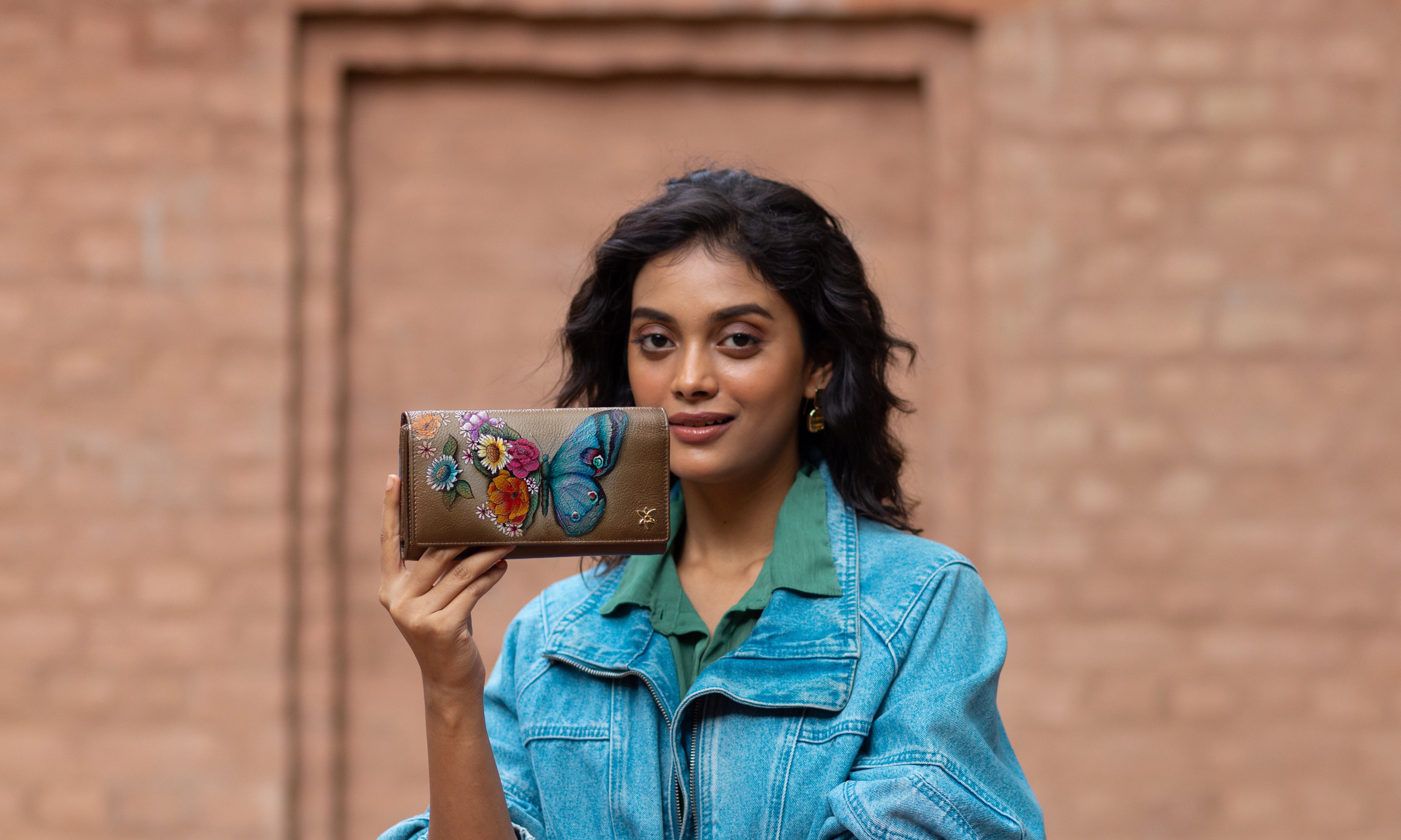 A woman in a pink sweater holds up a white wristlet wallet with a blue peacock design while standing outdoors in a courtyard area.