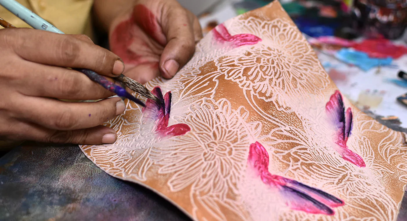 Person painting a floral design with red and black strokes on a piece of paper with intricate patterns, using a small brush.