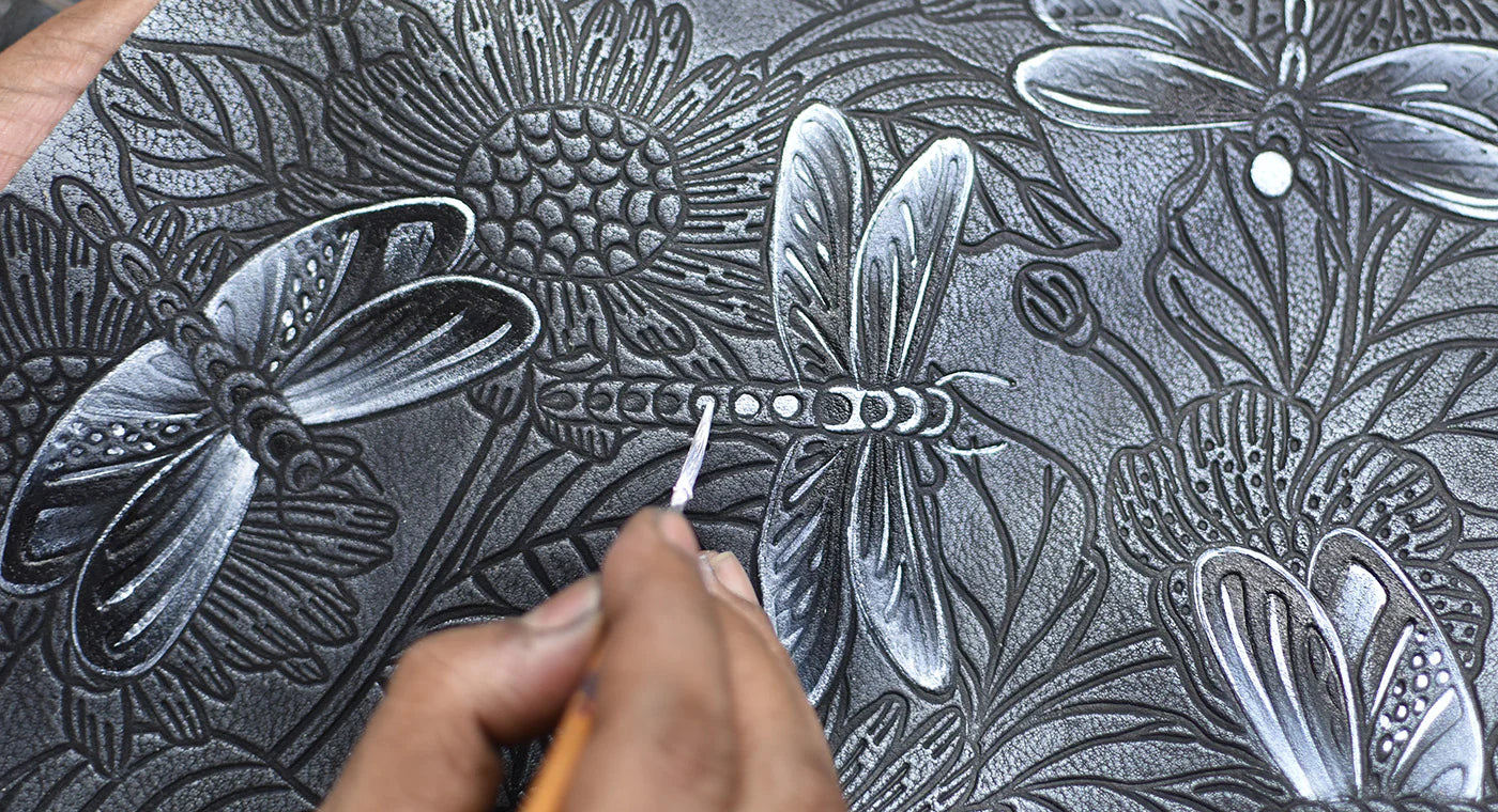 A person's hand is etching a detailed design of dragonflies and flowers into a black surface with a fine tool.