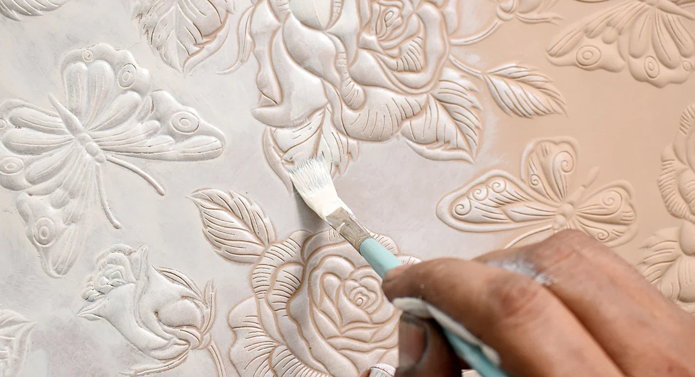 A hand holding a brush is painting over embossed floral and butterfly patterns on a surface.