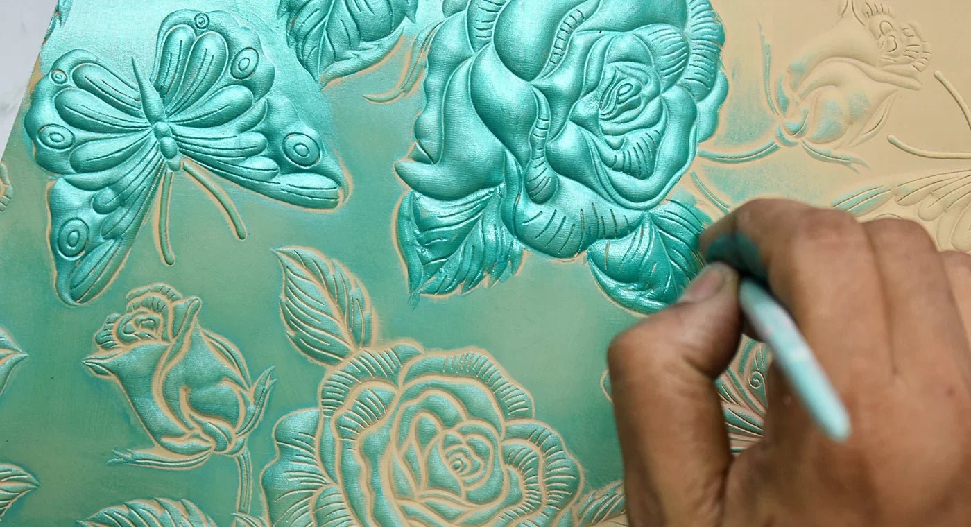 A close-up of someone's hand painting a light blue floral and butterfly design on a textured, embossed surface.