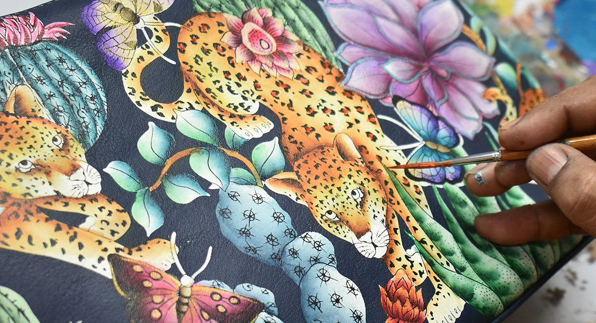 A hand is meticulously painting colorful, detailed images of leopards, flowers, and butterflies on a dark background canvas.