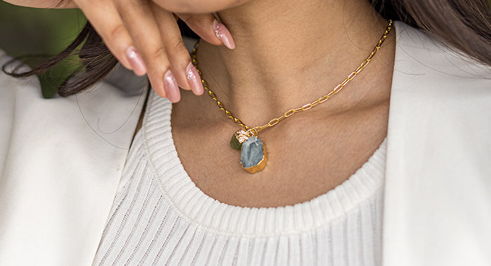 Close-up of a woman wearing a gold necklace with a large, oval aquamarine pendant, against a white top.