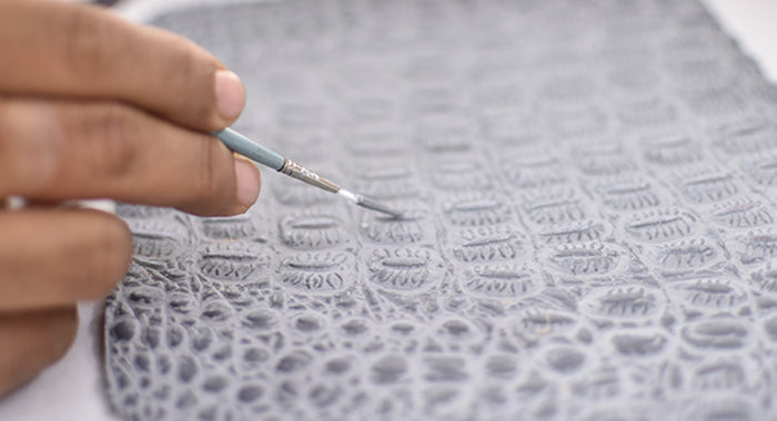 A hand uses a small brush to paint intricate patterns on a textured, grey surface.