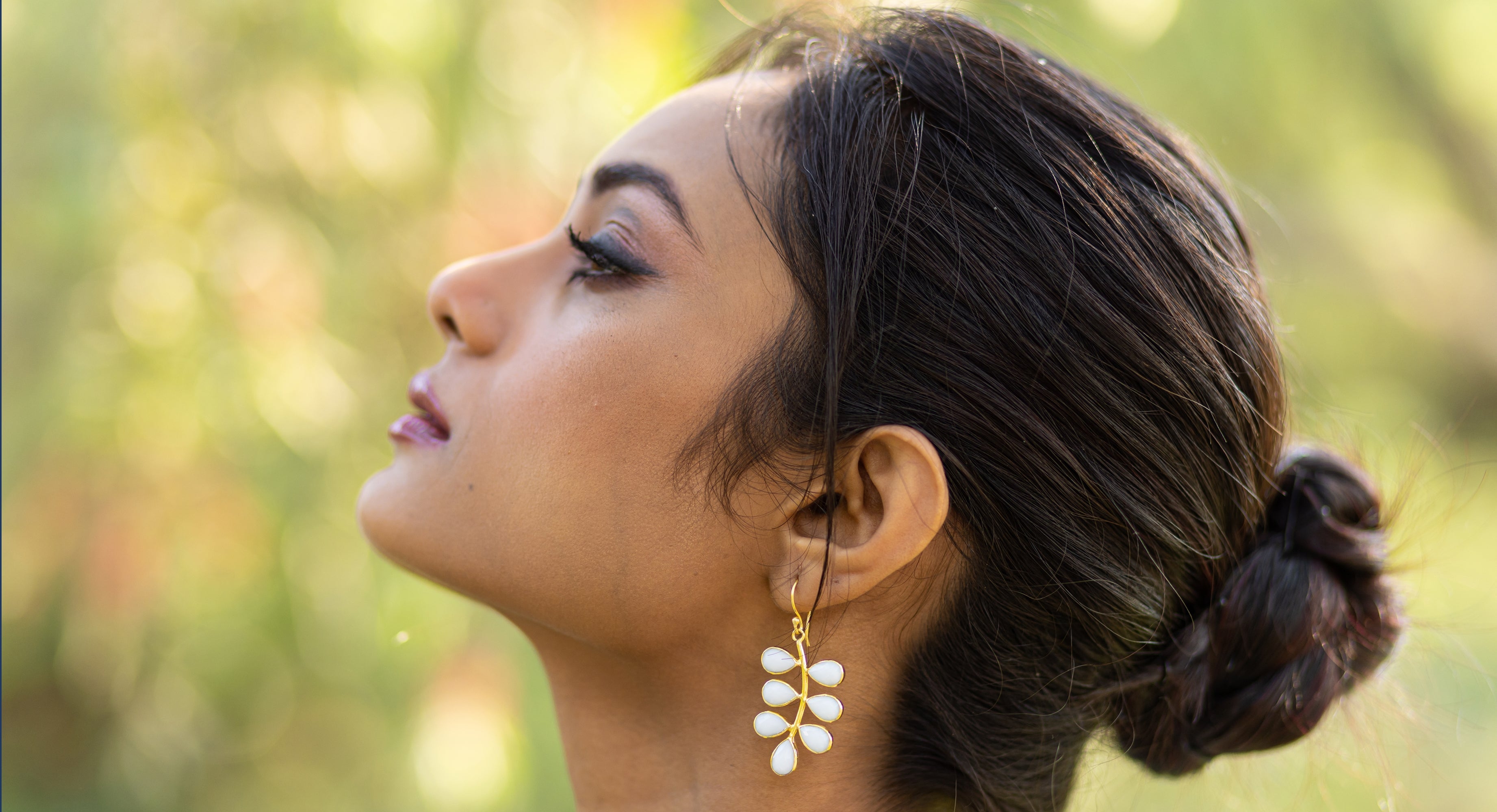 Profile of a person with dark hair in a bun, wearing white dangling earrings, looking ahead with a blurred green background.